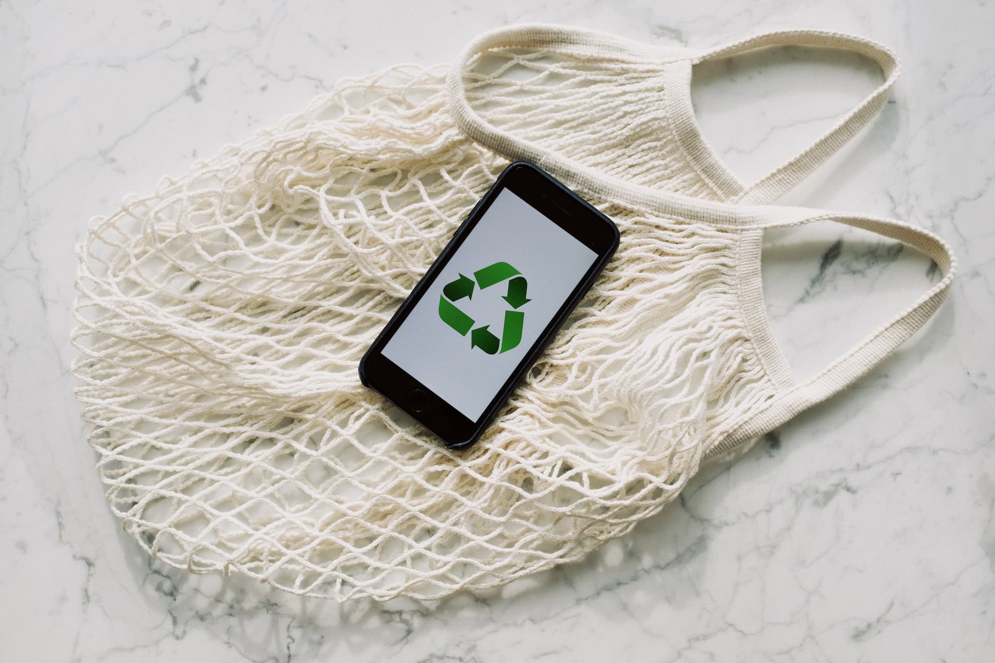 borsa riutilizzabile in rete, sotto a un telefono cellulare con simbolo riciclo cellulare con simbolo riciclo sopra a una borsa riutilizzabile in rete